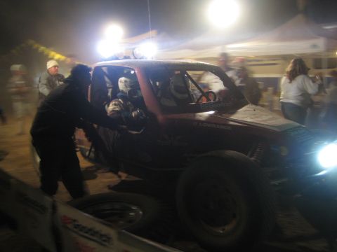 1990 Ford Bronco - pit crew member leans into a red off-road race truck during a nighttime pit stop under bright tent lighting.