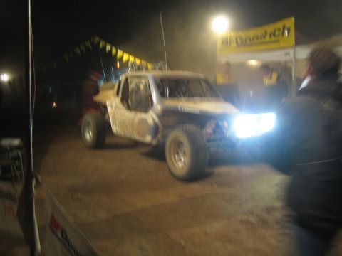 1990 Ford Bronco - white off-road race truck with bright headlights sits near a yellow BFGoodrich banner during a nighttime event.