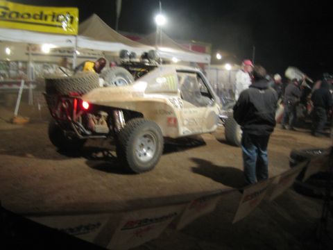 1990 Ford Bronco - rear of a tan off-road race truck idles in a crowded nighttime pit area as crew members stand nearby.