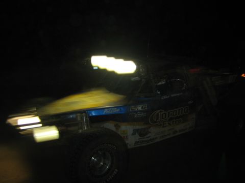 1990 Ford Bronco - trophy truck wearing Corona Extra and BFGoodrich livery streaks past with motion blur during a nighttime desert race.