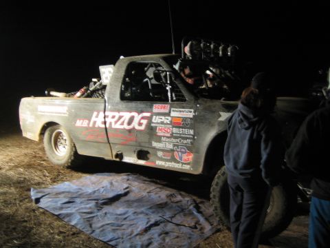 1990 Ford Bronco - off-road race truck lettered HERZOG and covered in sponsor decals sits on a tarp at night as a person stands beside it.