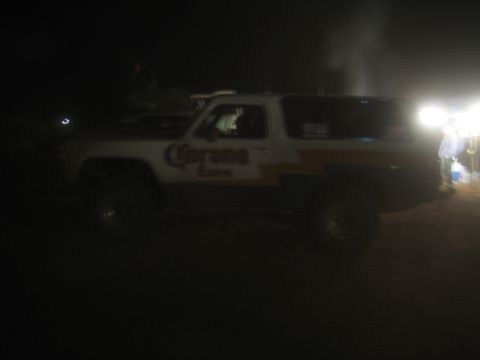 1990 Ford Bronco - lifted two-door SUV with Corona Extra livery sits in a dark, dusty lot with bright lights glaring behind it.