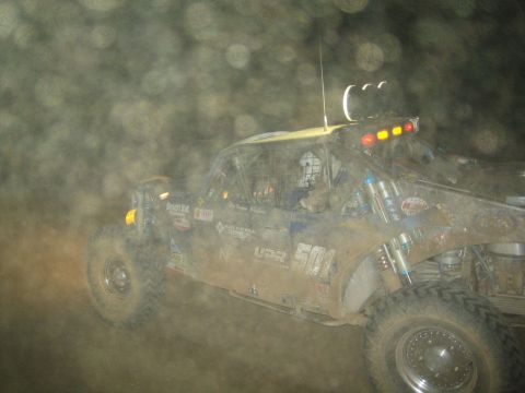 1990 Ford Bronco - off-road race buggy numbered 504 with a row of roof lights kicks up a heavy cloud of dust at night.