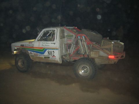 1990 Ford Bronco - white off-road race truck numbered 802 with an exposed rear tube chassis catches air during a nighttime race.