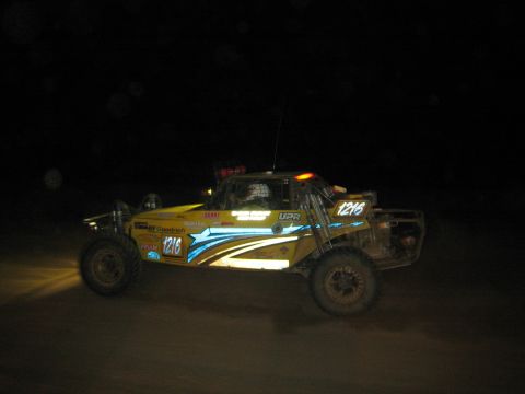 1990 Ford Bronco - yellow and blue off-road race buggy numbered 1216 with roof lights and SCORE decals races at night.