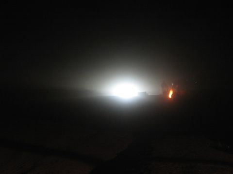 1990 Ford Bronco - bright white light source cuts through near-total darkness in a nighttime outdoor scene, with a small orange flame glowing at right.