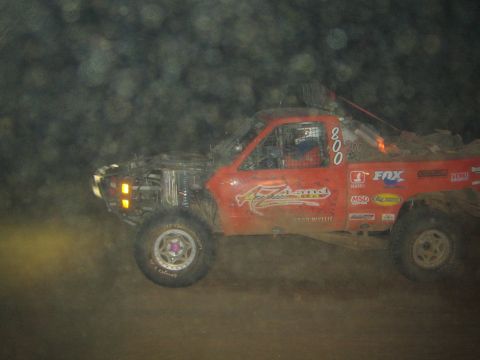 1990 Ford Bronco - red off-road race truck marked 800 with FOX and Method sponsor decals kicks up dust while racing at night.