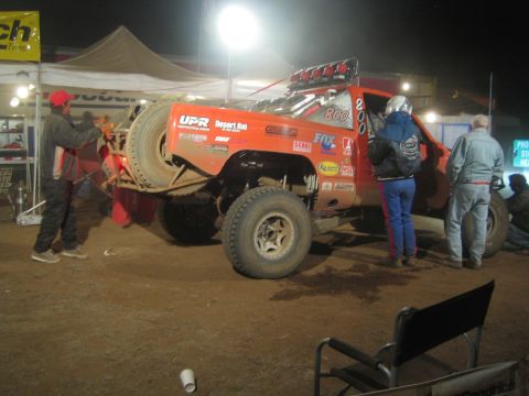 1990 Ford Bronco - orange off-road race truck numbered 800 sits raised on a jack in a lit pit area at night as crew members work around it.