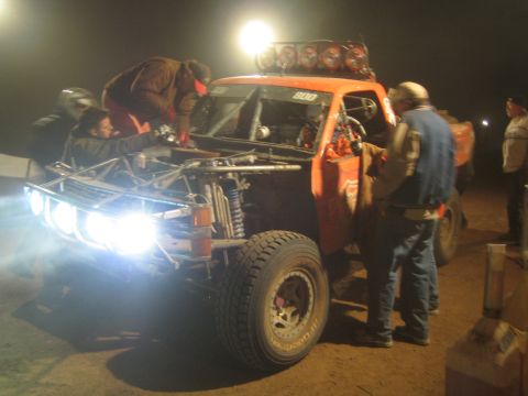 1990 Ford Bronco - Crew members work on an orange off-road race truck numbered 800 with its headlights and roof light bar illuminated during a night pit stop.