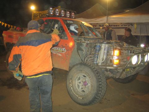 1990 Ford Bronco - man in an orange and blue jacket stands beside a mud-caked orange race truck numbered 800 under BFGoodrich banners at a night pit.