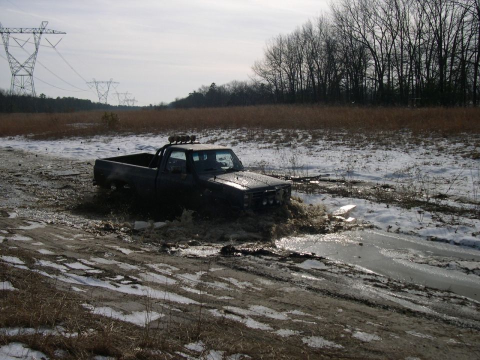 Dark short-bed pickup with roof light bar sitting in a muddy snow-patched field