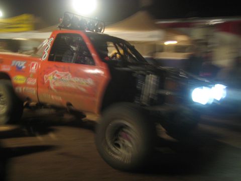 1990 Ford Bronco - orange off-road race truck with a roof light bar speeds through a lit pit area at night, captured with motion blur.