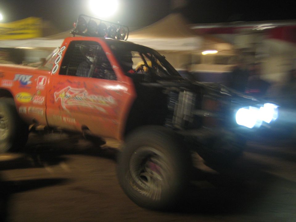 Orange race truck driving fast through a lit pit area at night