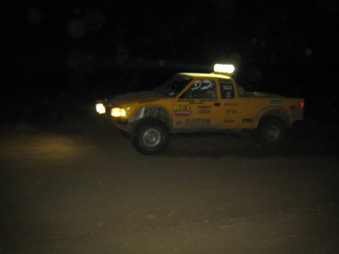 1990 Ford Bronco - yellow off-road race truck with a glowing roof light bar and Bilstein decals drives across dark terrain at night.