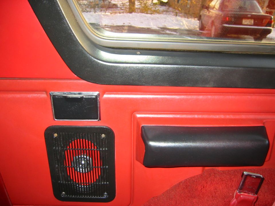 Red interior door panel with black armrest and speaker grille. A car is visible in the reflection of the window