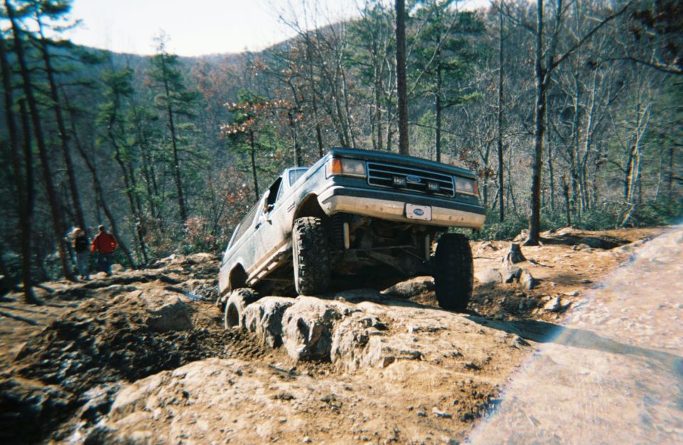 1990 Ford Bronco navigating a steep, rocky trail with trees in the background