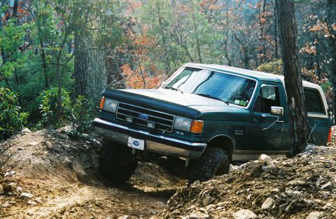 1990 Ford Bronco - A green Ford Bronco navigates a challenging off-road trail, with its front-left wheel deeply embedded in a dirt rut.