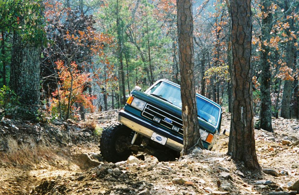 Green 1990 Ford Bronco tilted on a muddy trail with trees around