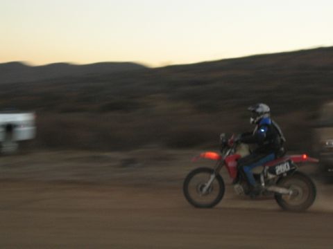 1990 Ford Bronco - helmeted rider on a red and blue dual-sport motorcycle numbered 260 rides across dirt terrain at dusk, captured with motion blur.