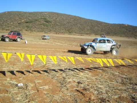 Light blue racing Baja Bug speeding across desert dirt past yellow flag line, number 576