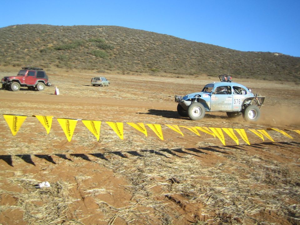 Light blue racing Baja Bug speeding across desert dirt past yellow flag line, number 576