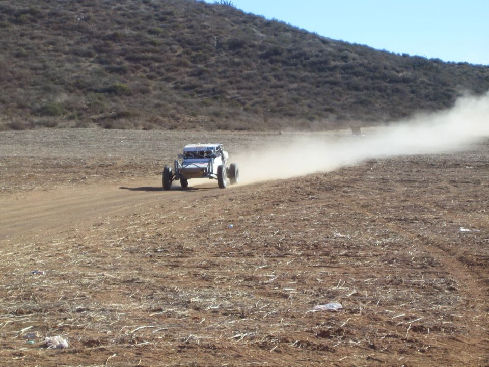 Open-frame off-road buggy racing away on dirt trail trailing a long dust cloud