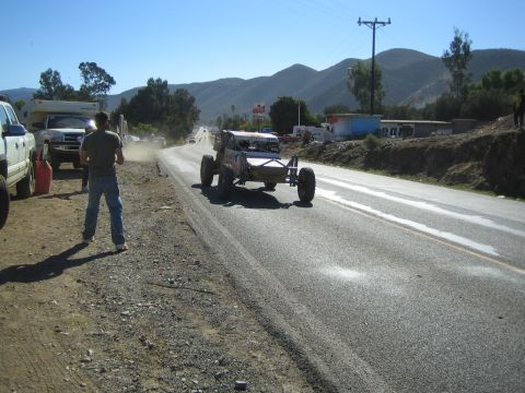 Open-frame race buggy on paved highway shoulder as spectators and trucks watch