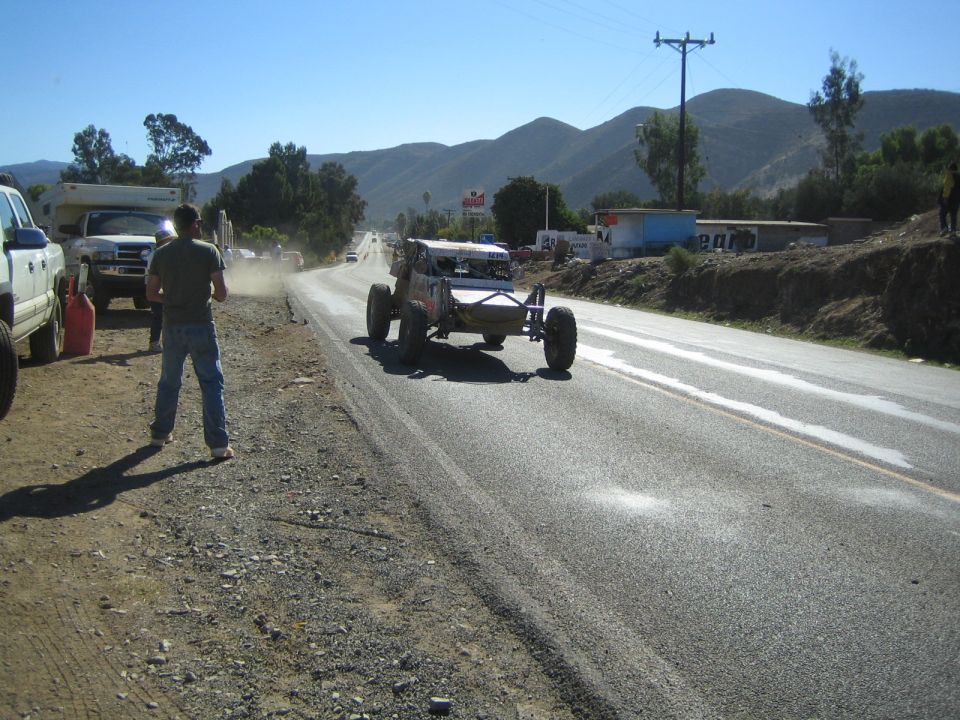 Open-frame race buggy on paved highway shoulder as spectators and trucks watch