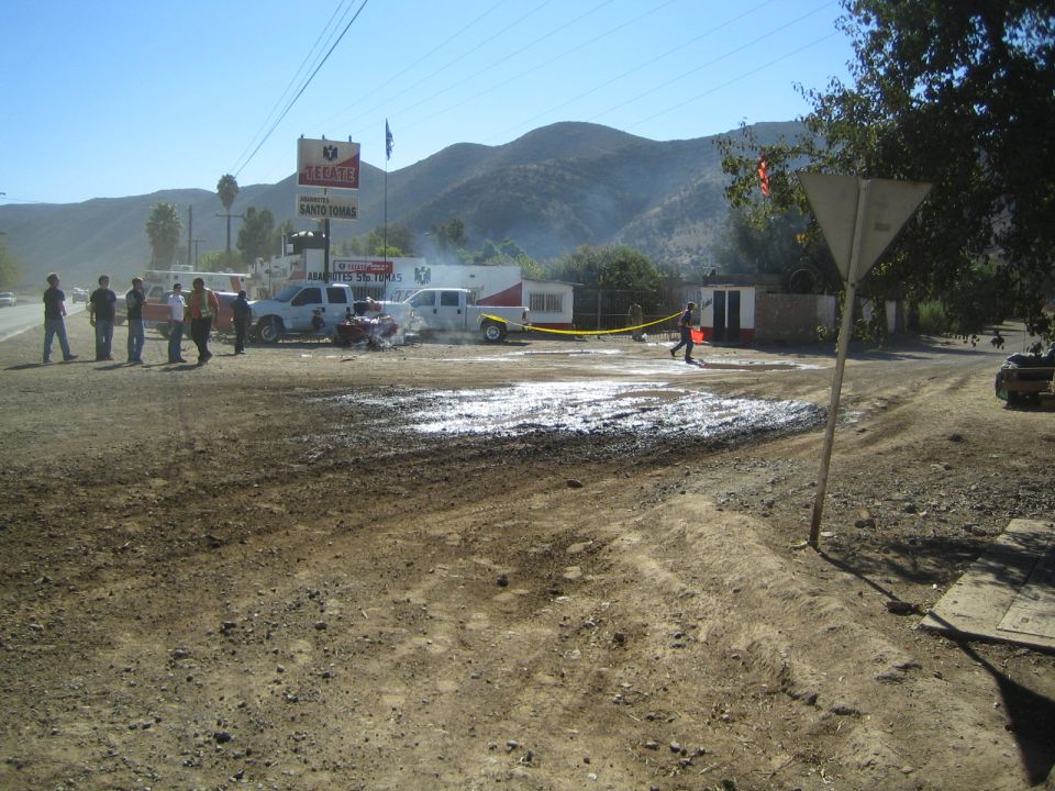 Dusty roadside scene with people, trucks and an ambulance near a Tecate-branded building
