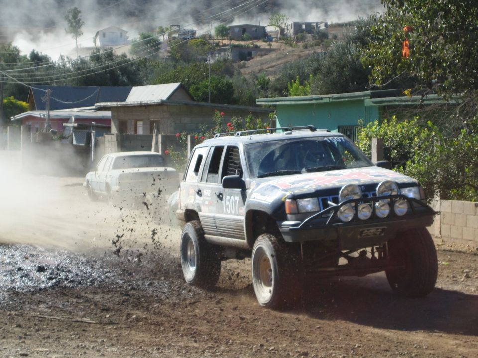 White Jeep Grand Cherokee race truck sliding through mud past village houses, number 1507