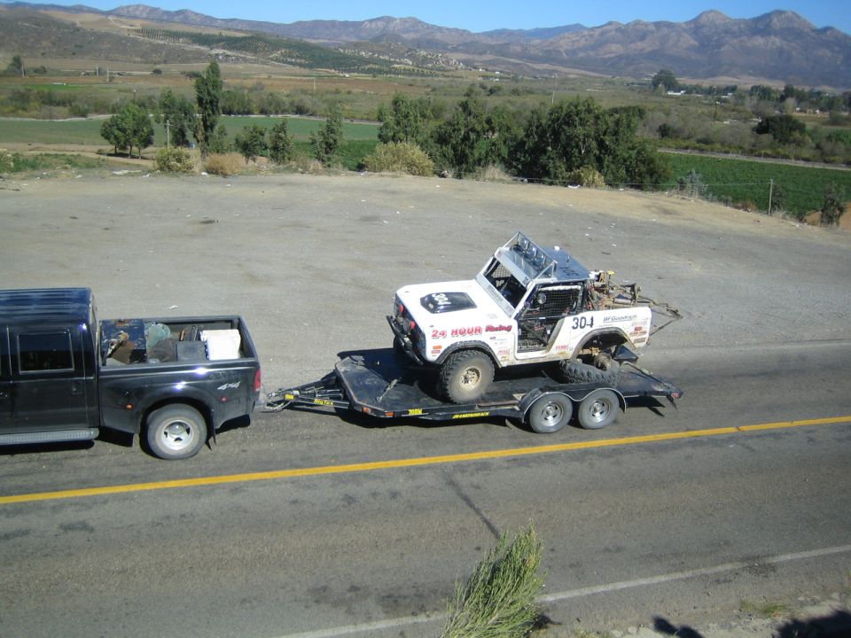 Black dually pickup towing a wrecked white Jeep race truck on a flatbed trailer