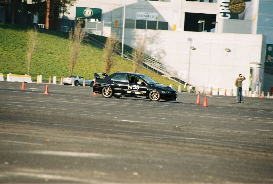Black Mitsubishi Lancer Evolution with number 55 on the door, driving on a track with orange cones. Another white car is visible in the background
