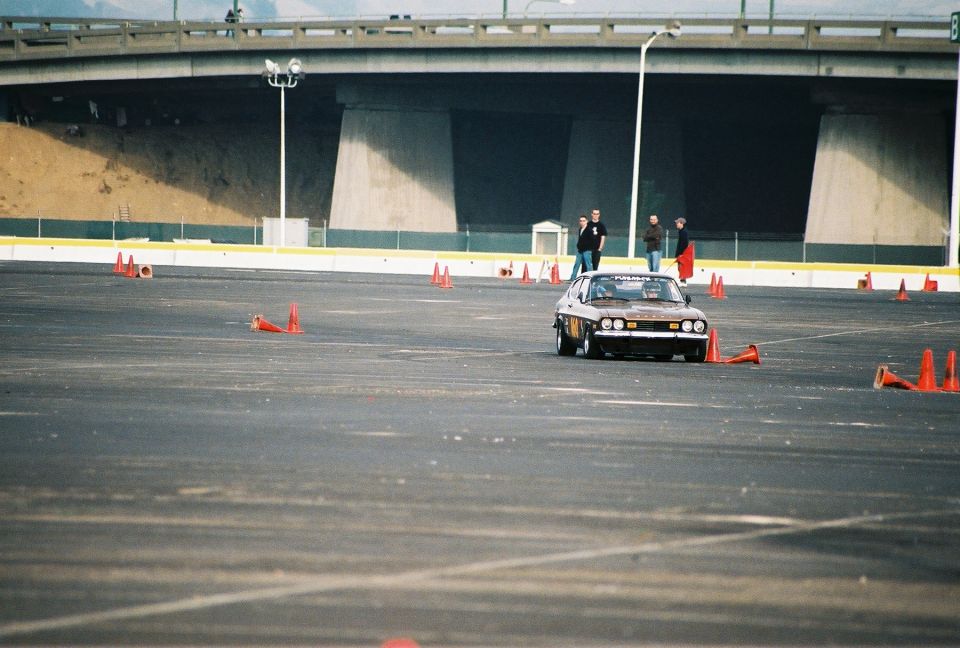 Brown sports car navigating an auto-cross course with orange cones. Supermotors.net logo in lower right