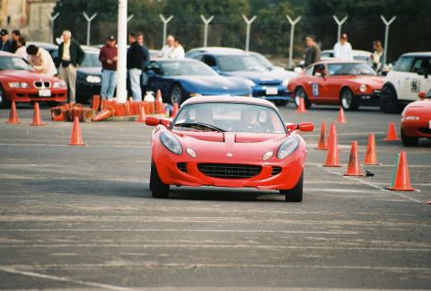 1997 Ford Probe - A red Lotus Elise navigates a course marked by orange cones, with other vehicles and spectators in the background.