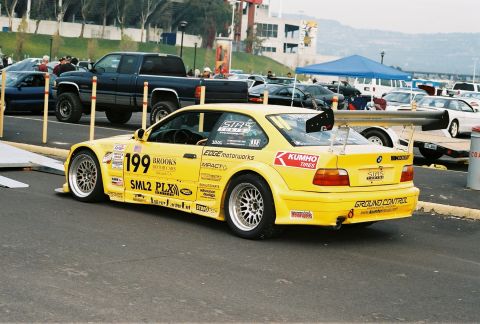 1997 Ford Probe - A yellow race car with numerous decals and a prominent rear spoiler is parked on asphalt, with other vehicles and tents in the background.
