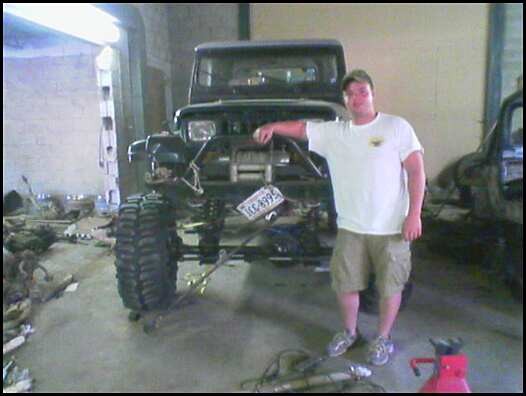 Man holding a license plate next to a lifted dark Jeep Wrangler under construction in a garage