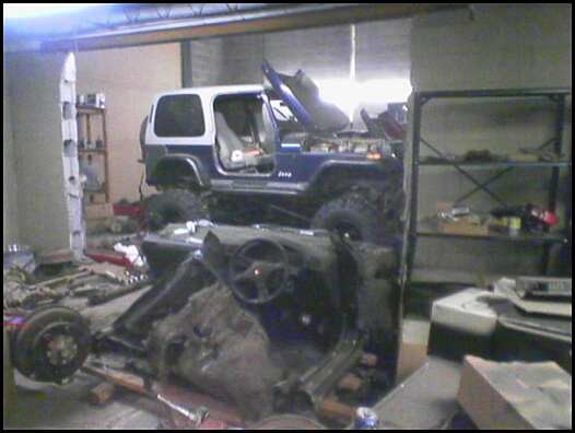 Blue Jeep Wrangler with hood up in garage behind a bare frame/chassis with steering wheel