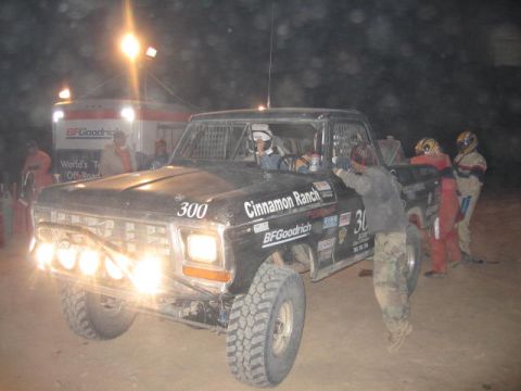 1990 Ford Bronco - black off-road race pickup numbered 300 and lettered Cinnamon Ranch sits under lights in a dusty night pit.