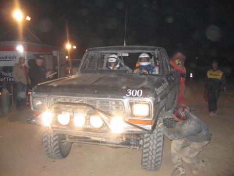 1990 Ford Bronco - black off-road race pickup numbered 300 with two helmeted occupants sits under bright lights at night.