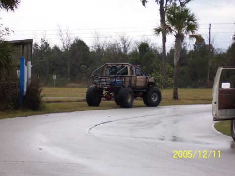 1989 Ford Bronco - A heavily modified off-road truck with a custom roll cage and massive tires is seen on a wet road, with another vehicle partially visible o…