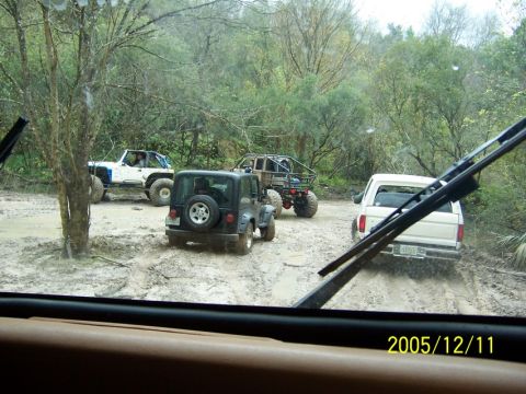 1989 Ford Bronco - Multiple off-road vehicles navigate a challenging muddy track in a wooded area.