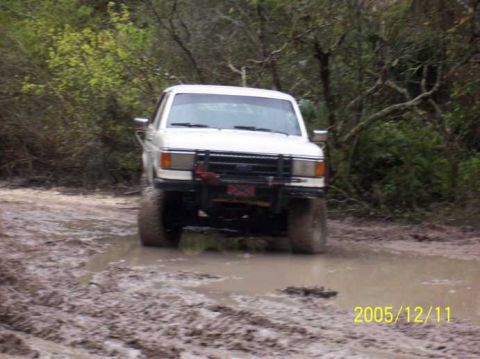 1989 Ford Bronco - A white 1989 Ford Bronco navigates a muddy trail, kicking up water as it goes.