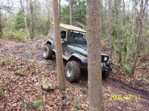 1989 Ford Bronco - A Jeep Wrangler navigates a muddy, wooded trail, kicking up dirt.