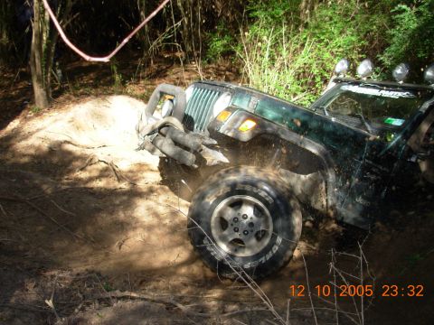 1987 Ford Bronco - A green Jeep Wrangler navigates a muddy off-road trail, with its front tire deep in the dirt.