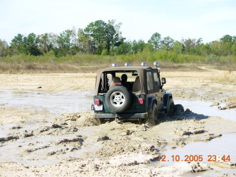 1987 Ford Bronco - A Jeep Wrangler navigates through a muddy terrain, kicking up water and dirt.