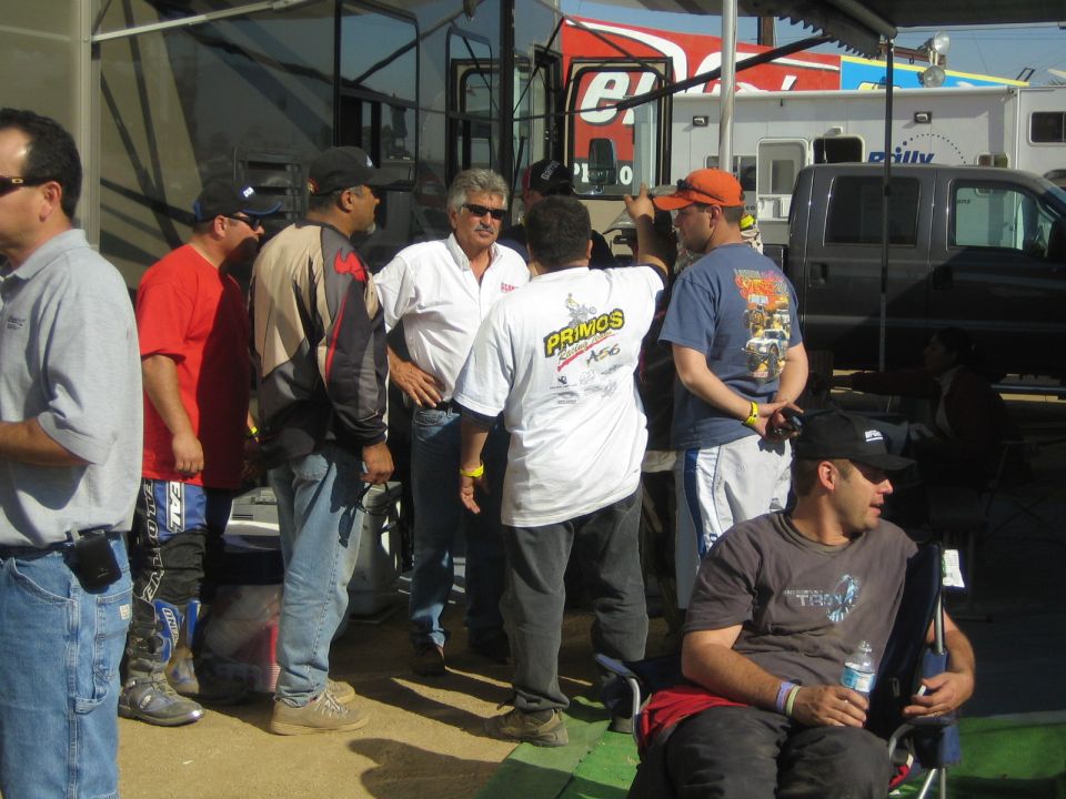 Group of men talking under a canopy beside an RV in a pit area