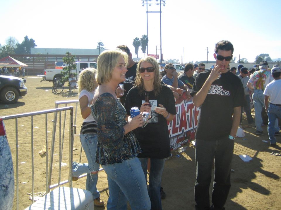 Three people standing at an outdoor event near a metal barricade in bright sun