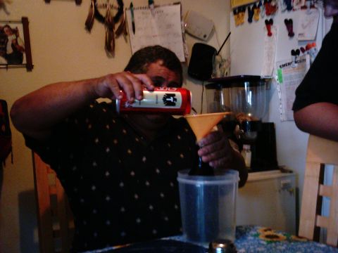 1986 Ford Bronco - A person is seen pouring a dark liquid from a red and white can into a clear plastic container using an orange funnel.