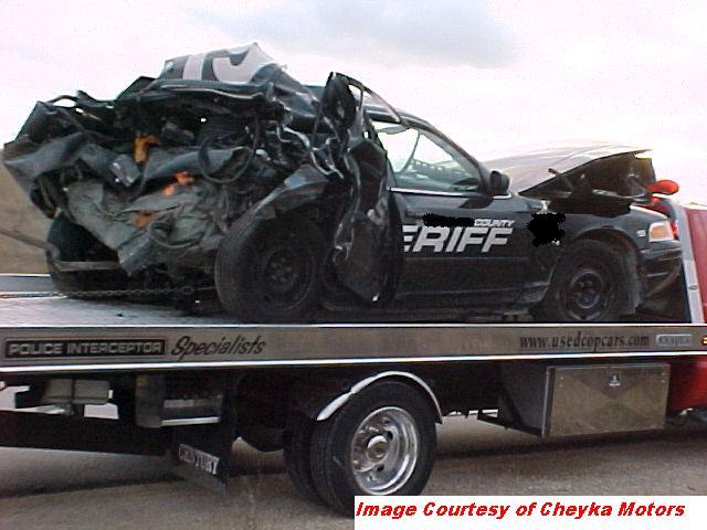 Severely damaged black sheriff's car with 'COUNTY SHERIFF' on the side, loaded onto a tow truck