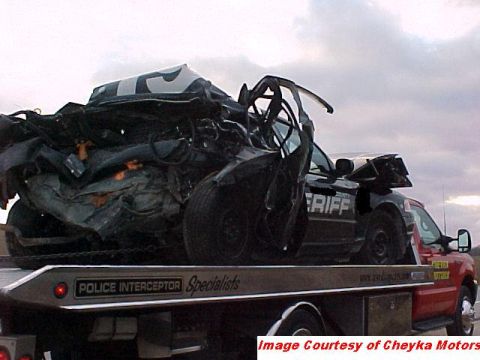 2000 Ford Crown Victoria - A severely damaged black police car, marked "SHERIFF", is loaded onto a red tow truck.